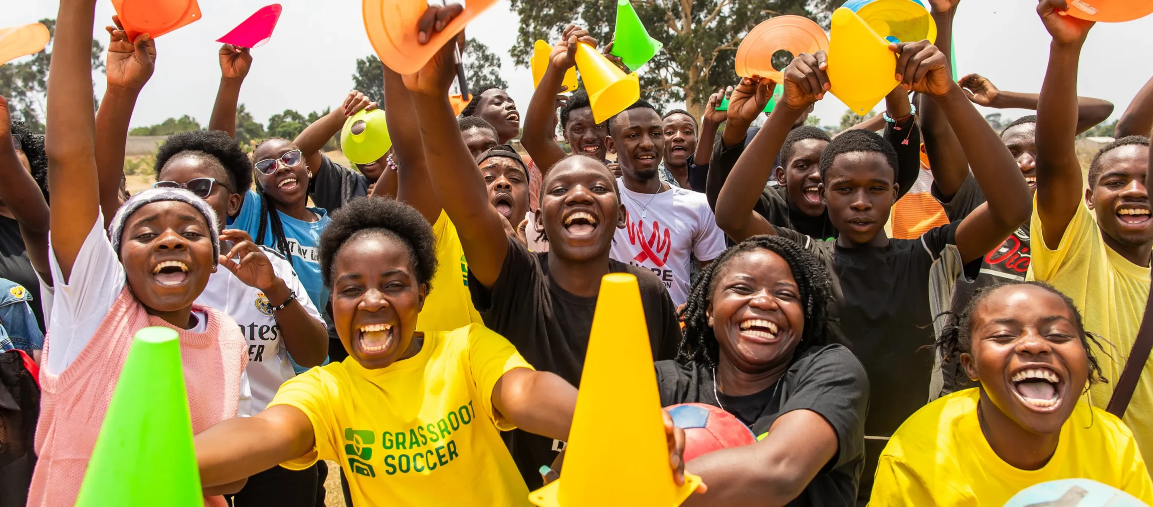 A Grassroot Soccer coach and a large group of young participants cheering and holding up colorful cones and equipment during an outdoor program session.