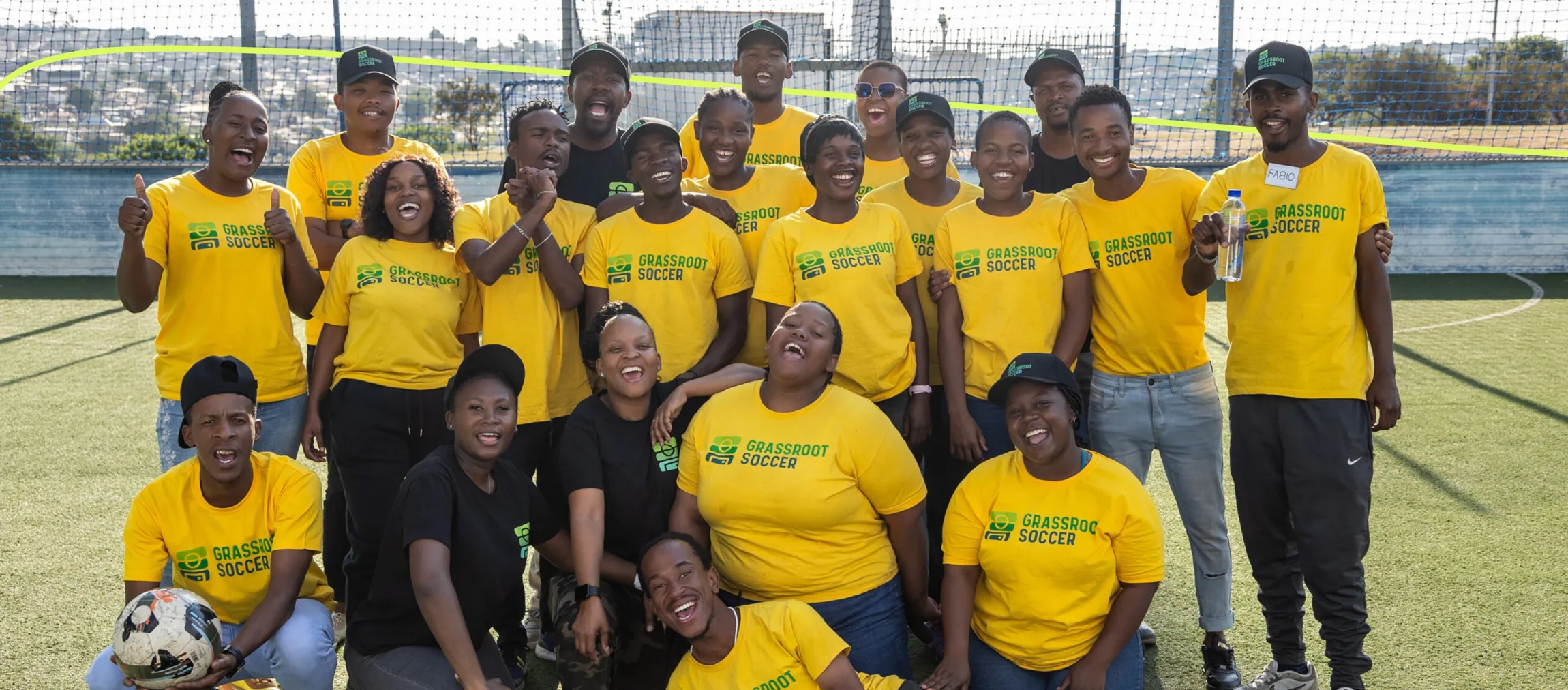 A large group of Grassroot Soccer coaches and staff posing together on a soccer field, wearing the new branded yellow t-shirts.